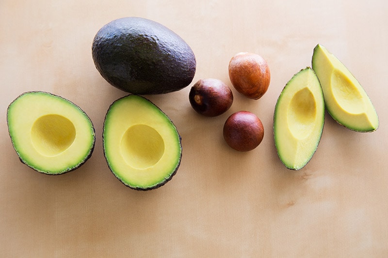 halved avocado next to a whole avocado, three pits, and two slices of  avocado