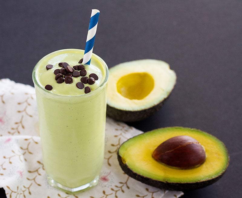 Glass of California Avocado milkshake topped with chocolate chips next to a cut avocado